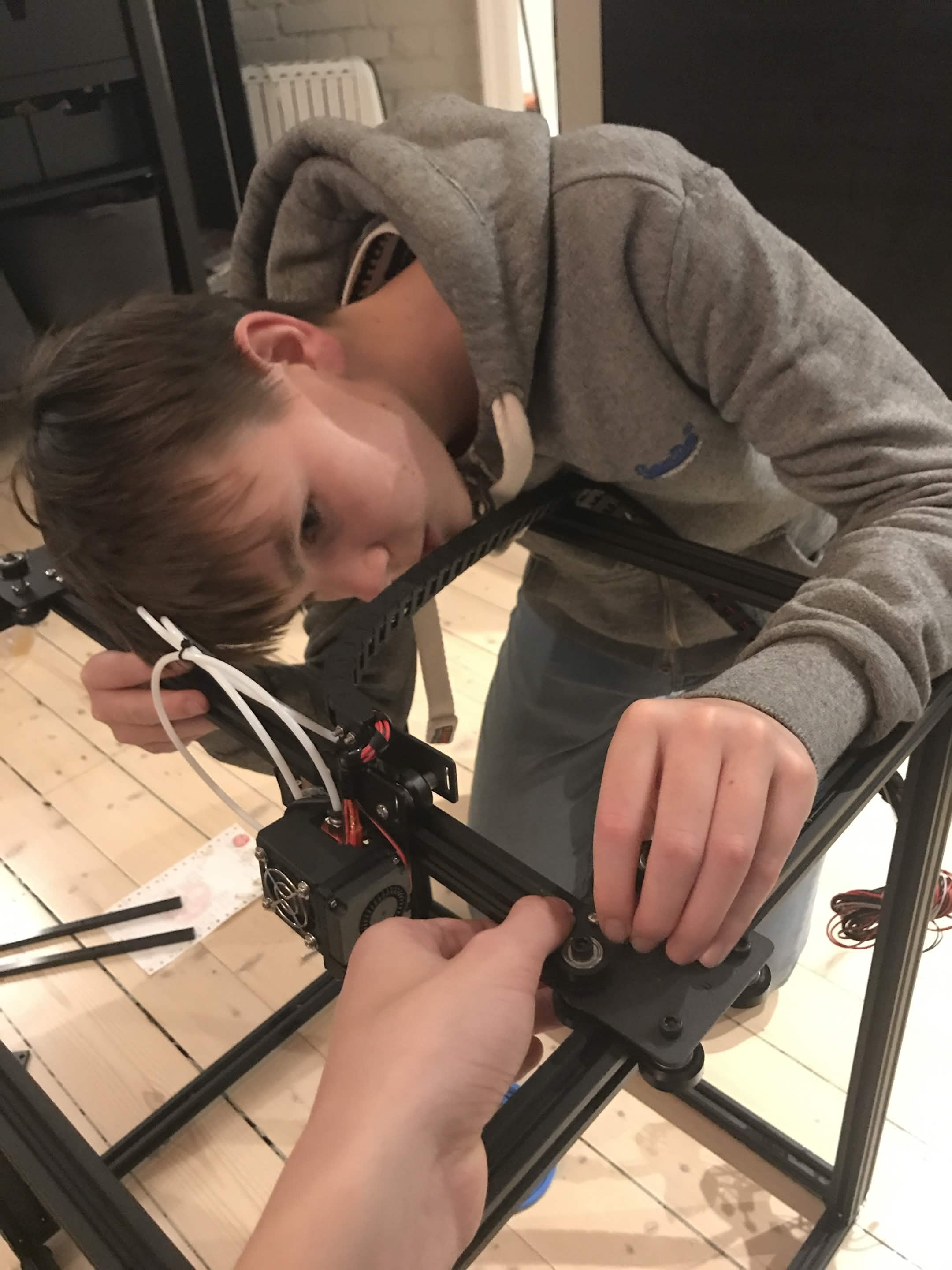 En tenåring lent over en aluminiumsprofilramme, strammer en reim på X-aksens vogn. Filamentekstruderen synes ved siden av.