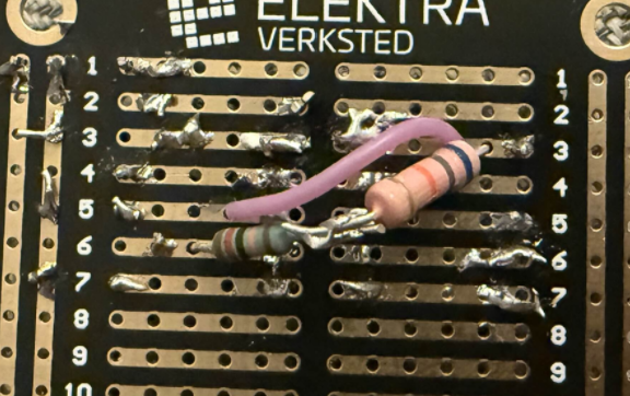 Nærbilde av et svart Elektra Verksted-veroboard med et par gjennomhullede motstander og en fiolett wire loddet mellom puter.